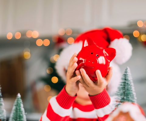 Un bambino davanti a delle decorazioni natalizie tiene in mano un salvadanaio rosso 