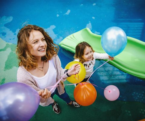 Mutter und kleines Mädchen mit Luftballons vor grüner Rutsche