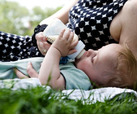 Kleinkind liegt mit Mutter im Gras und trinkt aus Babyfläschen 