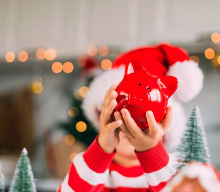Un bambino davanti a delle decorazioni natalizie tiene in mano un salvadanaio rosso 