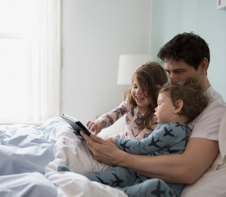 Vater mit zwei kleinen Kindern sitzen im Bett und spielen mit einem Tablet-PC