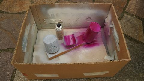 Mit Haftgrund und Farbe besprühte Dosen in einer Sprühkiste aus Karton