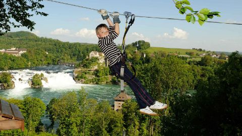 Junge rutscht die Seilrutsche hinunter, im Hintergrund sieht man den Rheinfall Junge rutscht die Seilrutsche hinunter, im Hintergrund sieht man den Rheinfall