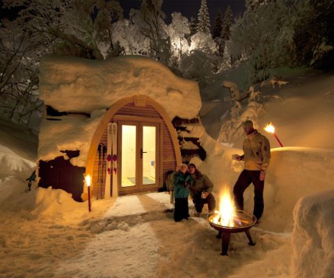 Familie vor verschneitem PODhouse bei Nacht im warmen Schein von Feuerschale und Fackeln
