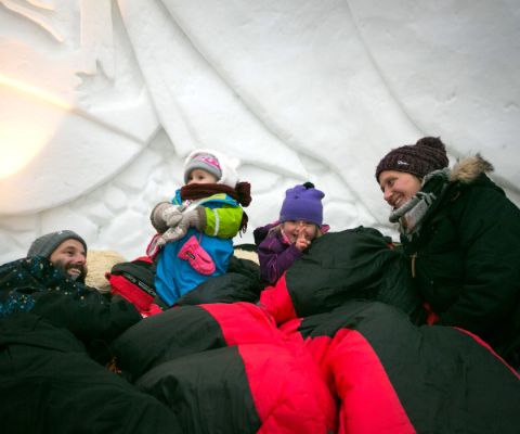 Familie mit Mützen, Handschuhen und Schlafsack übenachtet in einem Iglu