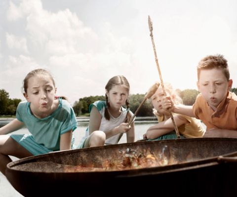 Quatre enfants tiennent des bâtons de bois entourés de pâte à pain et soufflent sur un brasero