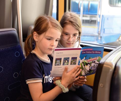 Zwei Mädchen in einem Zug lesen in der Ringo-Broschüre «Eine Statue für Rhina»