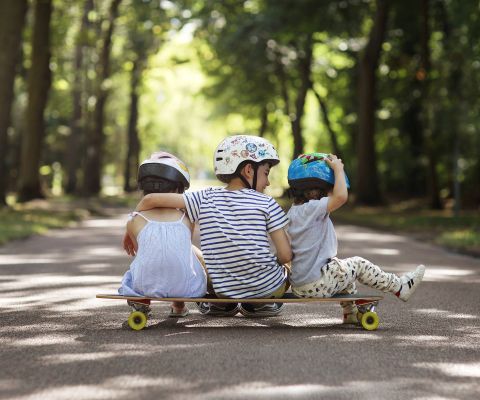 Drei Kinder mit Helm sitzen gemeinsam auf einem Skateboard