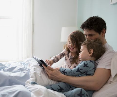 Un padre e due bambini piccoli seduti a letto che giocano con un tablet