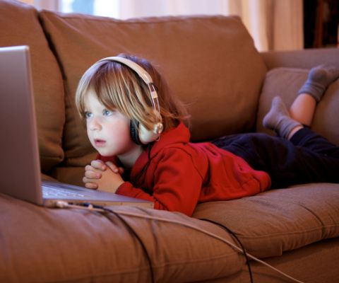 Un bambin, couché sur le canapé avec des écouteurs, regarde un ordinateur portable.