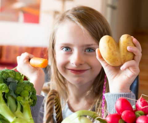 Mädchen zeigt lächelnd eine herzförmige Kartoffel und eine Möhre