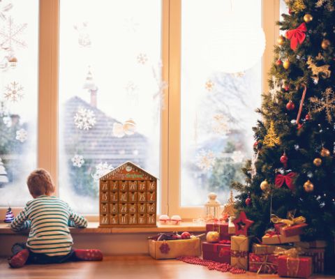 Junge schaut sitzend aus dem Fenster neben Adventskalender und Weihnachtsbaum