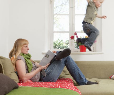 Un bambino si scatena saltando sul divano accanto alla mamma che legge