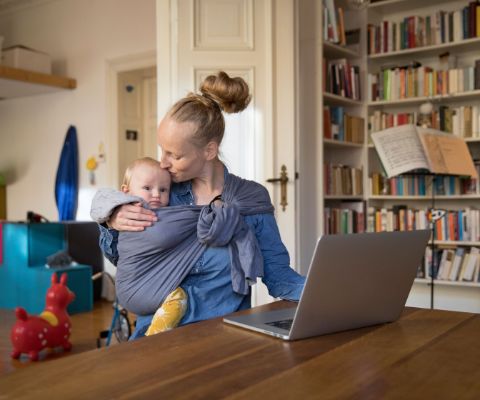 Maman portant son bébé dans une écharpe porte-bébé tandis qu’elle travaille sur l’ordinateur portable