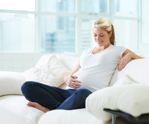 Une femme enceinte assise sur un canapé caresse son ventre en souriant.