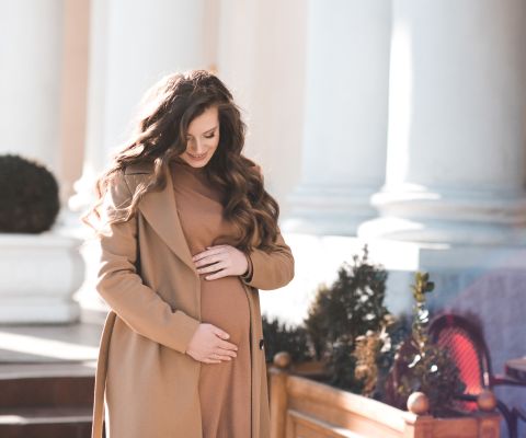 Donna incinta con mantello appoggia le mani sul pancione e sorride