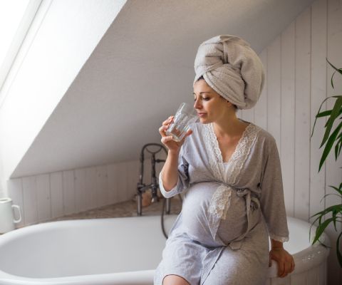 Schwangere sitzt auf dem Badewannenrand und trinkt ein Glas Wasser