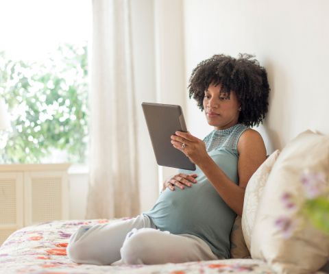 Donna incinta con la mano sul pancione legge sul tablet stando seduta a letto