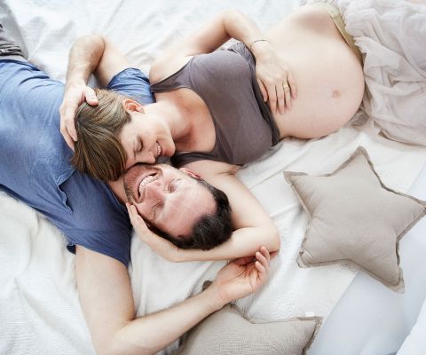 Une femme enceinte et un homme étendus sur un lit, blottis l’un contre l’autre, sourire aux lèvres