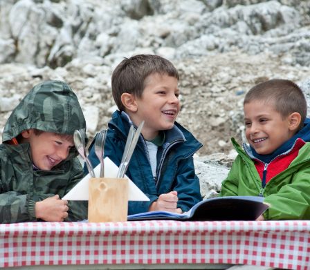 Tre ragazzini siedono sorridenti a un tavolo su una terrazza tra le montagne