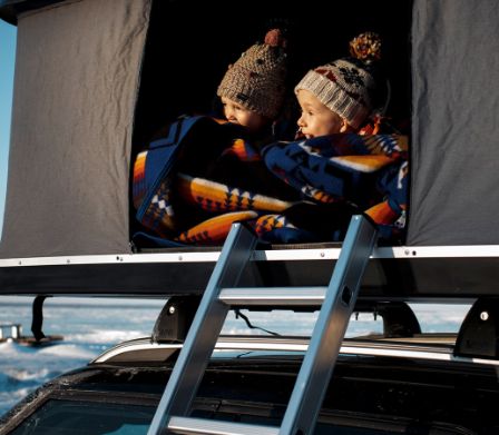 Zwei kleine Kinder mit Mützen schauen aus einem Camping-Dachzelt auf einem Auto