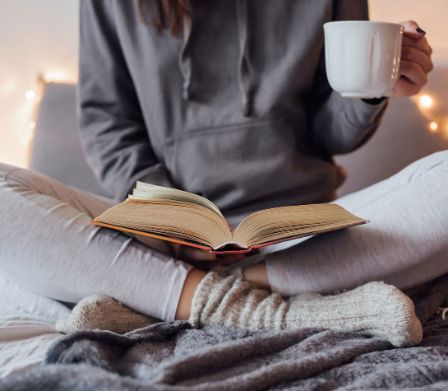 Una persona seduta a gambe incrociate legge un libro e tiene una tazza in mano 