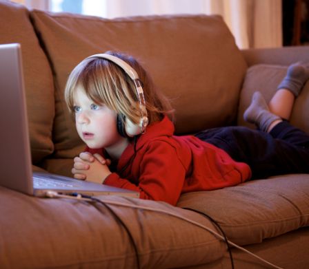Kleines Kind liegt mit Kopfhörer auf dem Sofa und schaut in einen Laptop