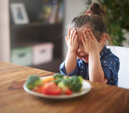 Une fillette se cache les yeux avec les mains, devant elle une assiette de légumes colorés