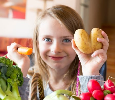 Mädchen zeigt lächelnd eine herzförmige Kartoffel und eine Möhre