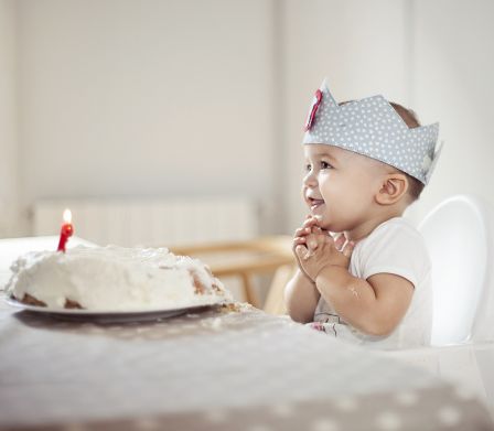 Baby sitzt im Kinderstuhl vor Geburtstagskuchen mit einer Kerze