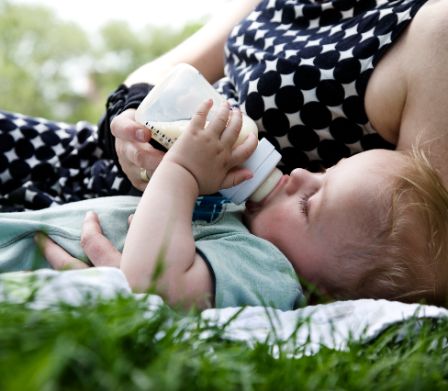 Kleinkind liegt mit Mutter im Gras und trinkt aus Babyfläschen 