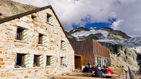 SAC Hütte mit Anbau und Gästen auf einer Terrasse